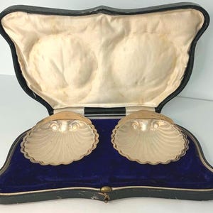 Könnte beinhalten: Ein Paar silberfarbene muschelförmige Schalen in einem schwarzen aufklappbaren Präsentationsetui. Das Innere des Etuis ist mit einem tiefblauen Samtboden und einem cremefarbenen Satinfutter mit zwei geformten Vertiefungen für die Schalen ausgestattet.