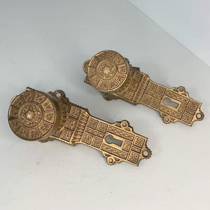 May include: Two antique brass door knobs and plates. The knobs are round with a detailed circular design. The plates have keyholes and rectangular patterns. The hardware is a warm gold color and appears to be vintage.