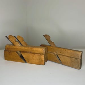 May include: Two antique wooden hand planes. The planes are made of light-colored wood with a rectangular body and a blade. The planes are positioned on a white surface.