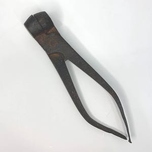 May include: A vintage, rusty metal tool with a wide, rectangular head and long, curved arms. The tool appears to be a type of pliers or clamp, likely used for a specific task. The metal has a weathered appearance, suggesting age and use.