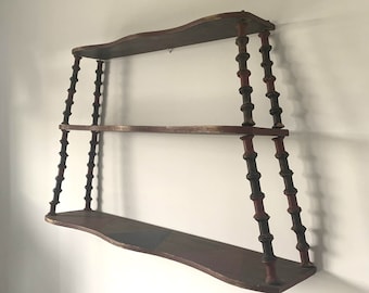 May include: A three-tiered wooden wall shelf with a distressed finish. The shelves are a dark brown with red and gold accents. The supports are dark brown with red details, creating a vintage aesthetic.