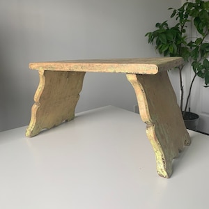 May include: A rustic wooden stool with a light green paint finish. The stool has a simple design with a flat top and curved legs.