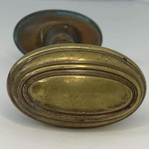 May include: An antique brass door knob with an oval shape and a ridged design. The knob has a tarnished, aged appearance, with hints of green oxidation on the back plate. The overall aesthetic suggests a vintage or retro style.