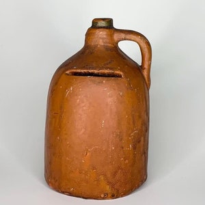 May include: A terracotta-colored ceramic jug with a handle and a slot in the front. The jug is a vintage-style piggy bank.