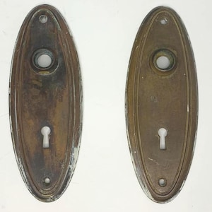 Könnte beinhalten: Zwei ovale antike Türschilder mit Schlüssellöchern und Schraubenlöchern. Die Schilder bestehen aus angelaufenem Messing mit dekorativem Rand. Die Schilder weisen Alters- und Gebrauchsspuren auf.