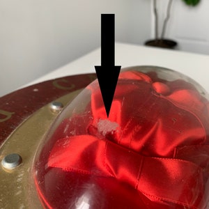 Peut inclure: Un ruban de satin rouge est pr&eacute;sent&eacute; sous un d&ocirc;me en verre transparent sur une base circulaire dor&eacute;e. La base pr&eacute;sente un motif rouge et or avec le mot "City" visible.