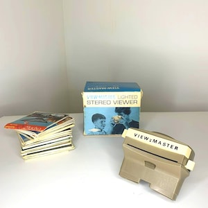 Könnte beinhalten: Ein Vintage View-Master-Set mit einem beigefarbenen Betrachter und einer blauen Box. Auf der Box steht "View-Master Lighted Stereo Viewer" und zeigt ein Bild von zwei Kindern. Ein Stapel View-Master-Rollen ist ebenfalls vorhanden.