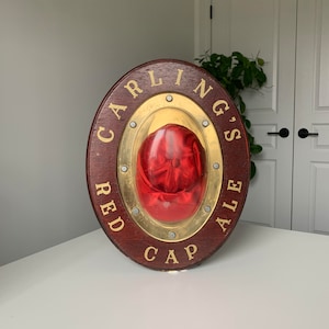 Peut inclure: Une enseigne ronde en bois avec un cadre en m&eacute;tal dor&eacute; et un bouchon rouge &agrave; l'int&eacute;rieur. L'enseigne porte l'inscription "Carling's Red Cap Ale".