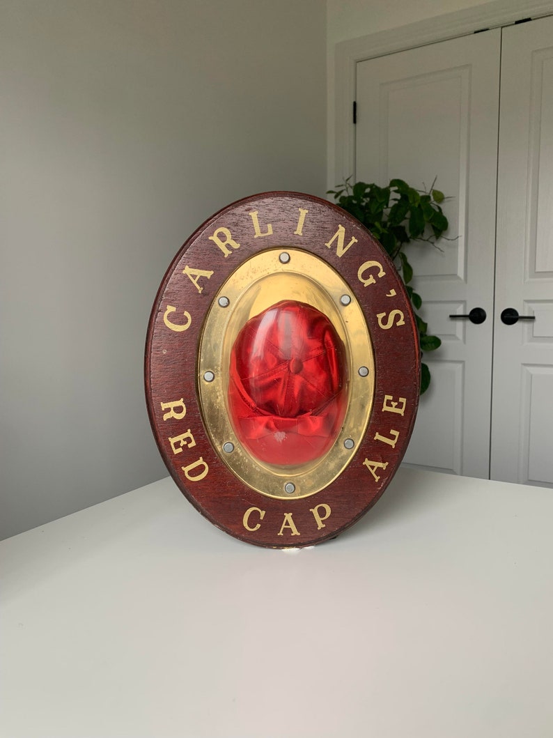 Peut inclure: Une enseigne ronde en bois avec un cadre en m&eacute;tal dor&eacute; et un bouchon rouge &agrave; l'int&eacute;rieur. L'enseigne porte l'inscription "Carling's Red Cap Ale".