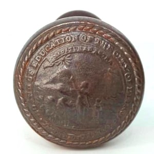 May include: A bronze-colored, round door knob with the text "US EDUCATION OFFICE" encircling a central emblem. The emblem features a detailed relief of figures and a banner. The knob has a rope-like border.