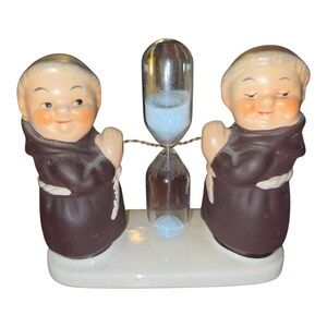 May include: A vintage hourglass with a clear glass center and blue sand, flanked by two ceramic figures of monks in brown robes. The base is white, and the figures have rosy cheeks and smiling expressions.