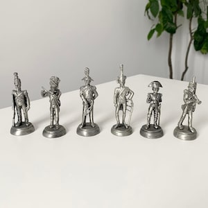 May include: Six pewter figurines of soldiers in various poses, wearing military uniforms and holding weapons. The figurines are displayed on a white surface.