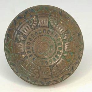 May include: A circular, antique bronze door knob with an intricate, embossed design. The knob features a central circular pattern surrounded by geometric shapes and floral accents. The bronze has a weathered patina with hints of green.