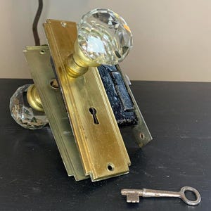 Op de afbeelding: Een vintage messing deurknop met een frontplaat en een heldere glazen kristallen knop. De knop is bevestigd aan een zwart metalen slot.