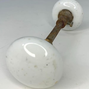 May include: A vintage door knob featuring two white, oval porcelain knobs joined by a rusty metal rod. The knobs have small, dark speckles and are secured with a screw.