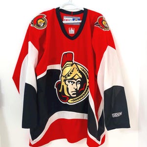 May include: Red, black, and white hockey jersey with a v-neck collar. The jersey features the Ottawa Senators logo on the front and shoulders. The sleeves are white with black accents and the CCM logo.