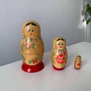 Può includere: Un set di tre bambole russe tradizionali a incastro, o bambole matrioska. La bambola più grande è dipinta con un motivo floreale e il viso di una donna. La bambola di mezzo è più piccola e ha un design simile. La bambola più piccola è la più semplice, con un design rosso e giallo.