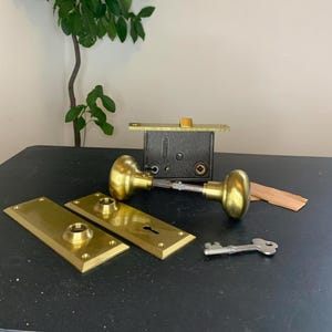 Peut inclure: Un ensemble de quincaillerie de porte en laiton antique, comprenant deux plaques de porte, deux boutons de porte, une serrure et une clé. La quincaillerie est en bon état et a une couleur dorée chaude.