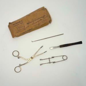 Könnte beinhalten: Eine Sammlung antiker chirurgischer Instrumente, darunter eine Schere, ein Skalpell und andere Werkzeuge. Die Werkzeuge sind auf einer weißen Oberfläche angeordnet, mit einer braunen Papierverpackung mit der Aufschrift "BURDIN'S CAPONIZING TOOLS" oben links.