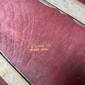 Peut inclure: Gros plan sur une valise en cuir rouge avec une sangle en tissu blanc. La valise porte une &eacute;tiquette dor&eacute;e en relief qui indique "J.A. Lamb Co. Detroit, Mich."