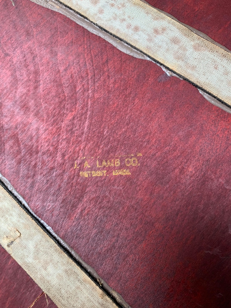 Peut inclure: Gros plan sur une valise en cuir rouge avec une sangle en tissu blanc. La valise porte une &eacute;tiquette dor&eacute;e en relief qui indique "J.A. Lamb Co. Detroit, Mich."
