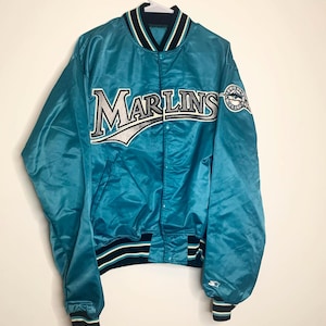 May include: A teal blue satin baseball jacket with the word "Marlins" embroidered on the front in silver and white lettering. The jacket has a black and white striped collar and cuffs.