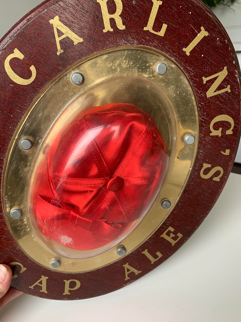 Peut inclure: Une enseigne en bois avec un cadre en m&eacute;tal dor&eacute; et une casquette de baseball en satin rouge &agrave; l'int&eacute;rieur. L'enseigne indique "Carling's Cap Ale".