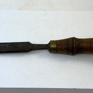 Peut inclure: Un ciseau vintage à manche en bois avec une virole en laiton et un biseau. Le ciseau est en métal et a une finition gris foncé.