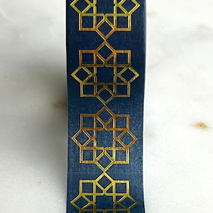 May include: A roll of blue washi tape with a gold geometric pattern. The pattern is a repeating design of a star with a square in the center.