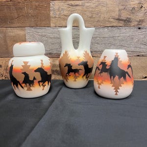 Könnte beinhalten: Drei weiße Keramikvasen mit einem schwarzen und orangefarbenen Pferdedesign. Die Vasen sind alle in verschiedenen Formen und Größen. Die größte Vase ist eine Dose mit Deckel. Die mittlere Vase ist eine traditionelle Vase im indianischen Stil mit Henkel. Die kleinste Vase ist eine runde Vase.