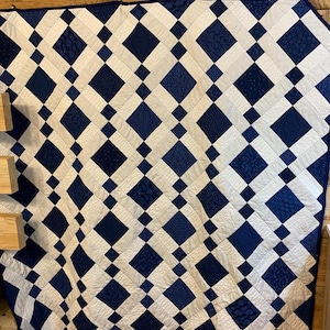 K&ouml;nnte beinhalten: Eine handgefertigte Steppdecke mit einem Rautenmuster in Marineblau und Wei&szlig;. Das Design der Steppdecke enth&auml;lt verschiedene Texturen und Muster innerhalb der blauen Rauten und erzeugt so ein visuelles Interesse. Die Steppdecke wird auf einer Holzoberfl&auml;che pr&auml;sentiert.