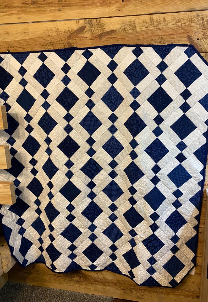 K&ouml;nnte beinhalten: Eine handgefertigte Steppdecke mit einem Rautenmuster in Marineblau und Wei&szlig;. Das Design der Steppdecke enth&auml;lt verschiedene Texturen und Muster innerhalb der blauen Rauten und erzeugt so ein visuelles Interesse. Die Steppdecke wird auf einer Holzoberfl&auml;che pr&auml;sentiert.