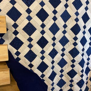 K&ouml;nnte beinhalten: Eine gesteppte Decke mit einem sich wiederholenden Rautenmuster in Marineblau und Off-White. Die Rauten sind in einem Raster angeordnet, mit abwechselnden Mustern aus einfarbigen Marineblau und gemusterten Stoffen. Die Kanten sind mit einem einfarbigen Marineblau-Rand versehen.