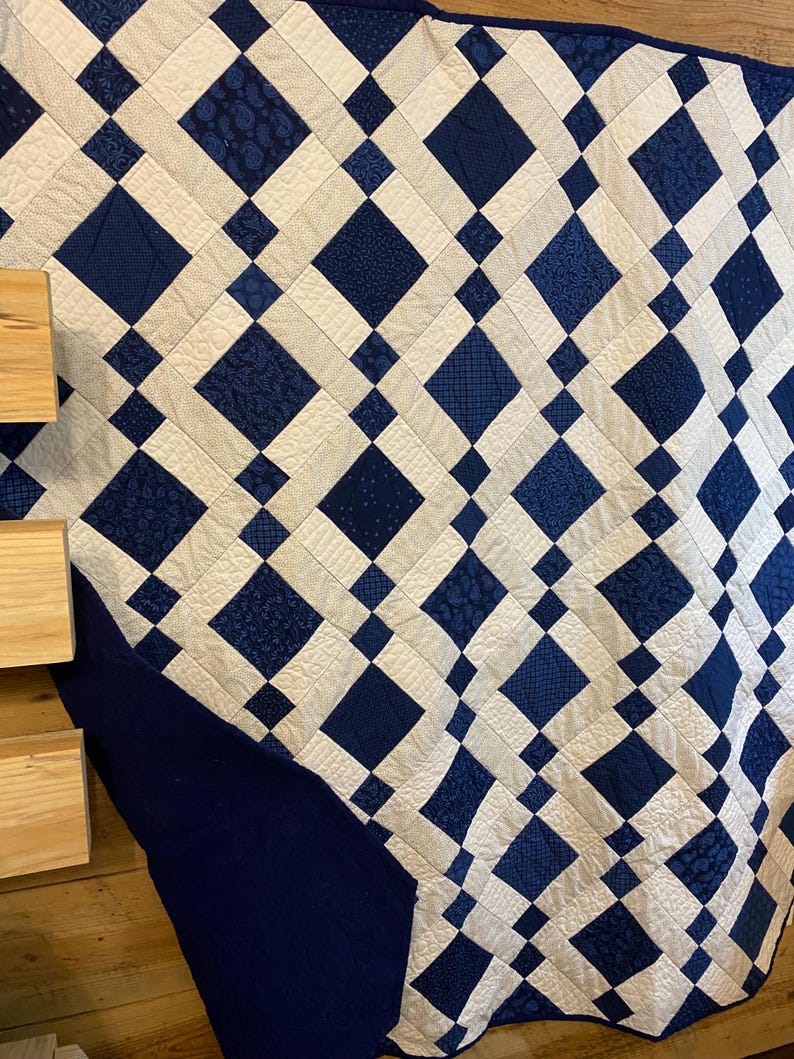 K&ouml;nnte beinhalten: Eine gesteppte Decke mit einem sich wiederholenden Rautenmuster in Marineblau und Off-White. Die Rauten sind in einem Raster angeordnet, mit abwechselnden Mustern aus einfarbigen Marineblau und gemusterten Stoffen. Die Kanten sind mit einem einfarbigen Marineblau-Rand versehen.