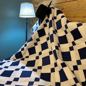 K&ouml;nnte beinhalten: Eine handgefertigte Steppdecke mit einem geometrischen Muster aus marineblauen und wei&szlig;en Quadraten. Die Steppdecke ist &uuml;ber einem h&ouml;lzernen Kopfteil drapiert, mit einem schwarzen Hut darauf. Im Hintergrund befindet sich eine Lampe mit beigem Schirm.