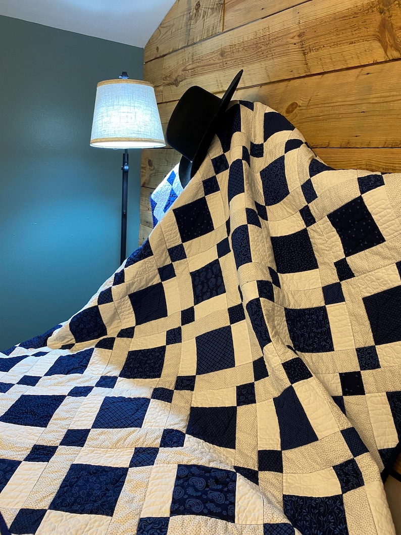 K&ouml;nnte beinhalten: Eine handgefertigte Steppdecke mit einem geometrischen Muster aus marineblauen und wei&szlig;en Quadraten. Die Steppdecke ist &uuml;ber einem h&ouml;lzernen Kopfteil drapiert, mit einem schwarzen Hut darauf. Im Hintergrund befindet sich eine Lampe mit beigem Schirm.