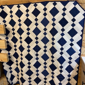 K&ouml;nnte beinhalten: Eine handgefertigte Steppdecke mit einem Rautenmuster in Marineblau und Wei&szlig;. Die Steppdecke besteht aus zusammengen&auml;hten Stoffquadraten, die ein geometrisches Design bilden. Die Steppdecke wird an einer Holzwand ausgestellt.