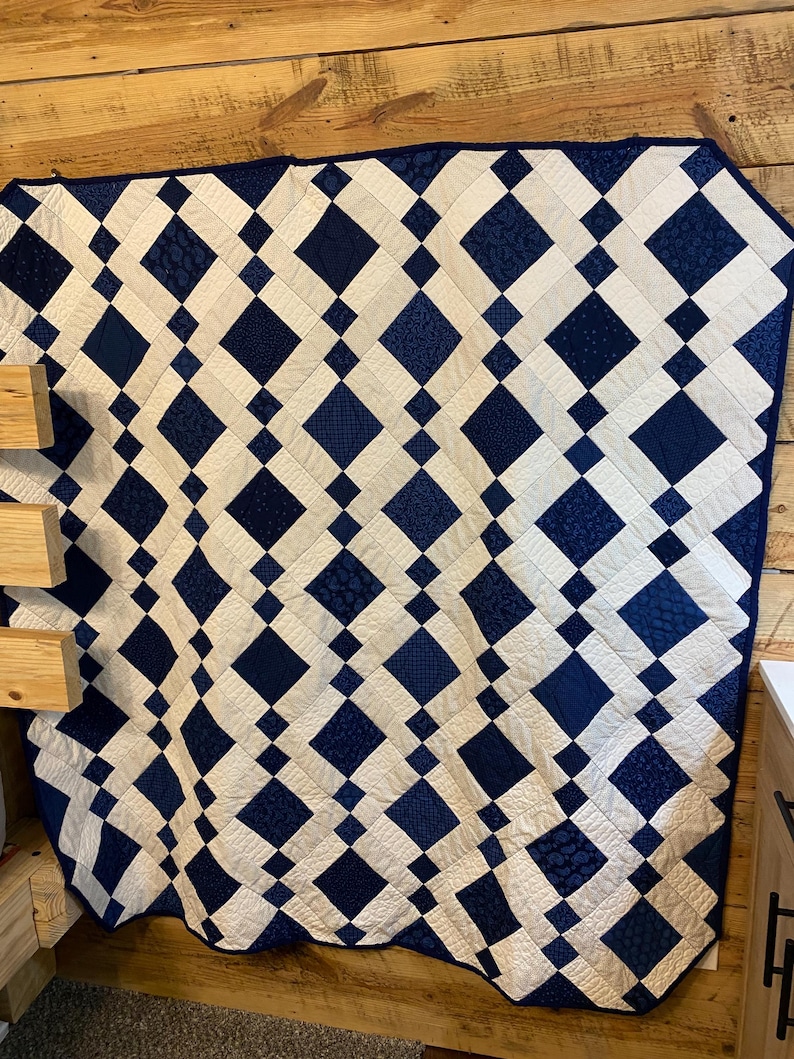 K&ouml;nnte beinhalten: Eine handgefertigte Steppdecke mit einem Rautenmuster in Marineblau und Wei&szlig;. Die Steppdecke besteht aus zusammengen&auml;hten Stoffquadraten, die ein geometrisches Design bilden. Die Steppdecke wird an einer Holzwand ausgestellt.