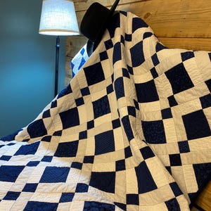 K&ouml;nnte beinhalten: Eine handgefertigte Steppdecke mit einem marineblauen und wei&szlig;en Schachbrettmuster. Die Steppdecke ist &uuml;ber einem h&ouml;lzernen Kopfteil drapiert, mit einem schwarzen Hut darauf. Das Design der Steppdecke zeigt verschiedene marineblaue Muster.