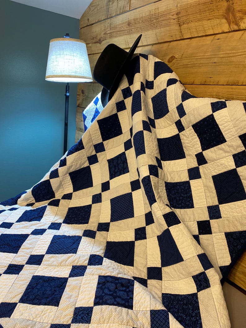 K&ouml;nnte beinhalten: Eine handgefertigte Steppdecke mit einem marineblauen und wei&szlig;en Schachbrettmuster. Die Steppdecke ist &uuml;ber einem h&ouml;lzernen Kopfteil drapiert, mit einem schwarzen Hut darauf. Das Design der Steppdecke zeigt verschiedene marineblaue Muster.