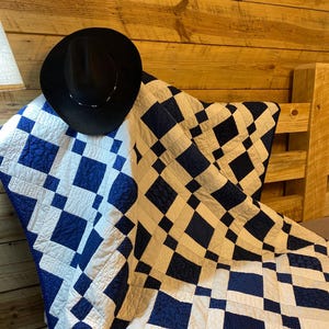 K&ouml;nnte beinhalten: Ein schwarzer Cowboyhut liegt auf einer blau-wei&szlig;en Steppdecke mit Rautenmuster. Die Steppdecke ist &uuml;ber einem h&ouml;lzernen Kopfteil drapiert. Das Design der Steppdecke zeigt abwechselnd blaue und wei&szlig;e Quadrate.