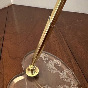 May include: A gold-colored pen and heart-shaped pen holder. The pen is inserted into the holder, which is clear with an etched floral design. The pen holder is made of clear acrylic. The pen and holder are on a wooden surface.