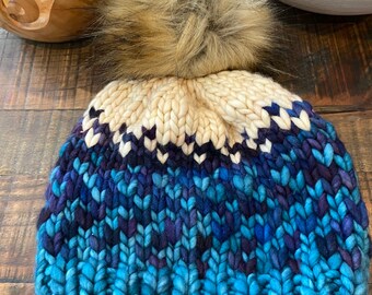 May include: A blue, purple, and white knitted beanie with a fluffy brown pom pom. The beanie has a chunky knit texture.
