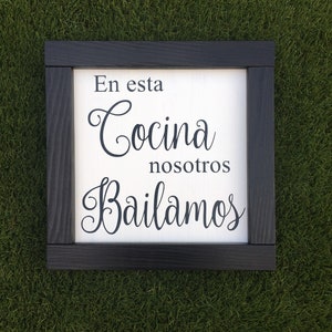 May include: A white wooden sign with black lettering that reads "En esta cocina nosotros bailamos" which translates to "In this kitchen we dance". The sign has a black wood frame.