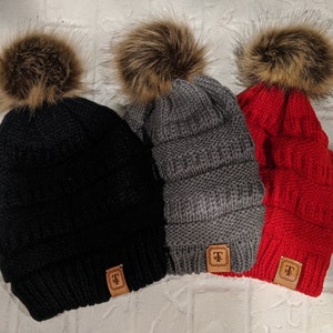 May include: Three knit winter hats with faux fur pom poms. The hats are black, gray, and red. Each hat has a brown leather tag with the letter "T" on it.