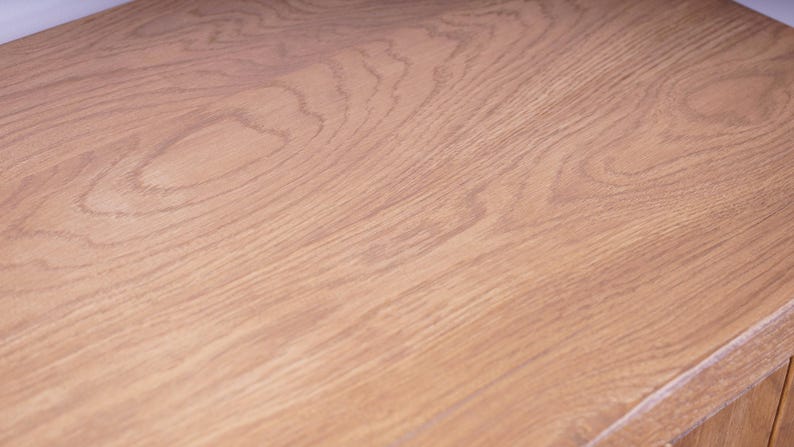 May include: A light brown wooden surface featuring a prominent natural grain pattern with swirling lines and organic textures. The wood displays a warm, tan color palette with visible knots and elongated fibers running across the flat plane.