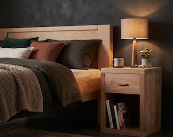 Puede incluir: Una estructura de cama de madera clara con cabecero, acompañada de una mesita de noche de madera a juego con un cajón y un estante abierto con libros. Una lámpara de mesa con pantalla cilíndrica de tela se sitúa sobre la mesita junto a una taza.