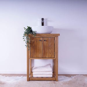 May include: A wooden bathroom vanity cabinet with two doors and a lower shelf holding folded white towels. A white ceramic vessel sink sits on top with a dark faucet. A small green plant hangs over the left side of the wooden vanity unit.