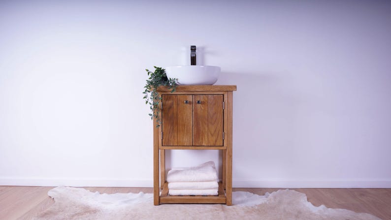 May include: A wooden bathroom vanity cabinet with two doors and a lower shelf holding folded white towels. A white ceramic vessel sink sits on top with a dark faucet. A small green plant hangs over the left side of the wooden vanity unit.