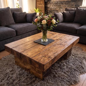 Puede incluir: Una mesa de centro rectangular construida con tablones de madera gruesos te&ntilde;idos de oscuro con un patr&oacute;n de veta visible. La mesa presenta una base baja de estilo bloque y sostiene un jarr&oacute;n de vidrio transparente lleno de un ramo de flores rosas, blancas y rojas.